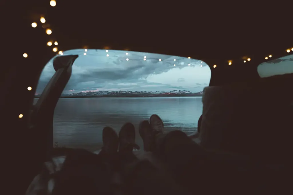 Cozy-Car-Camping Two people relaxing inside a campervan at dusk, looking out over a lake and mountains with string lights above