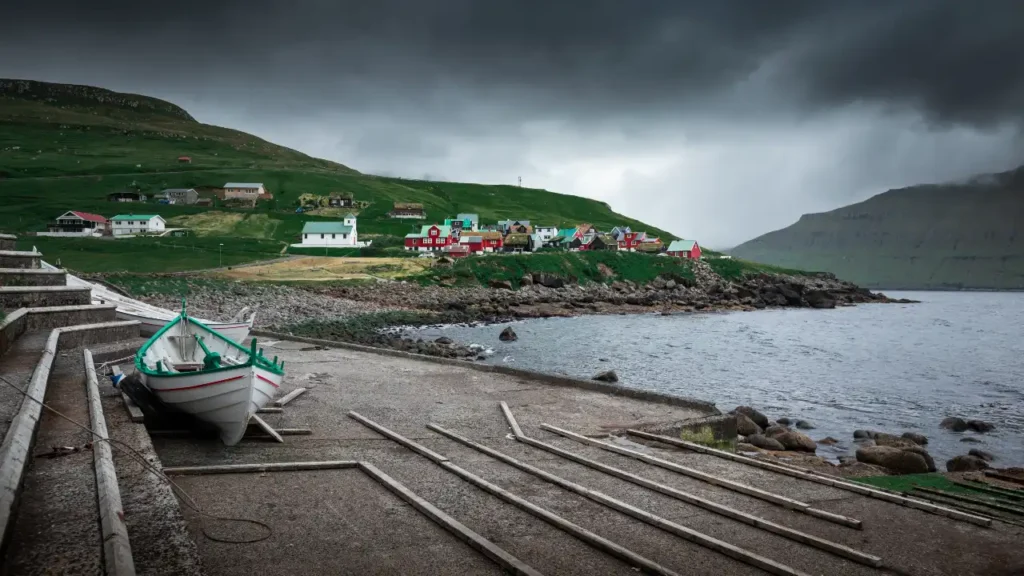 Small fishing boats pulled ashore with colorful hillside houses in Elduvík village, Faroe Islands, under dramatic cloudy skies