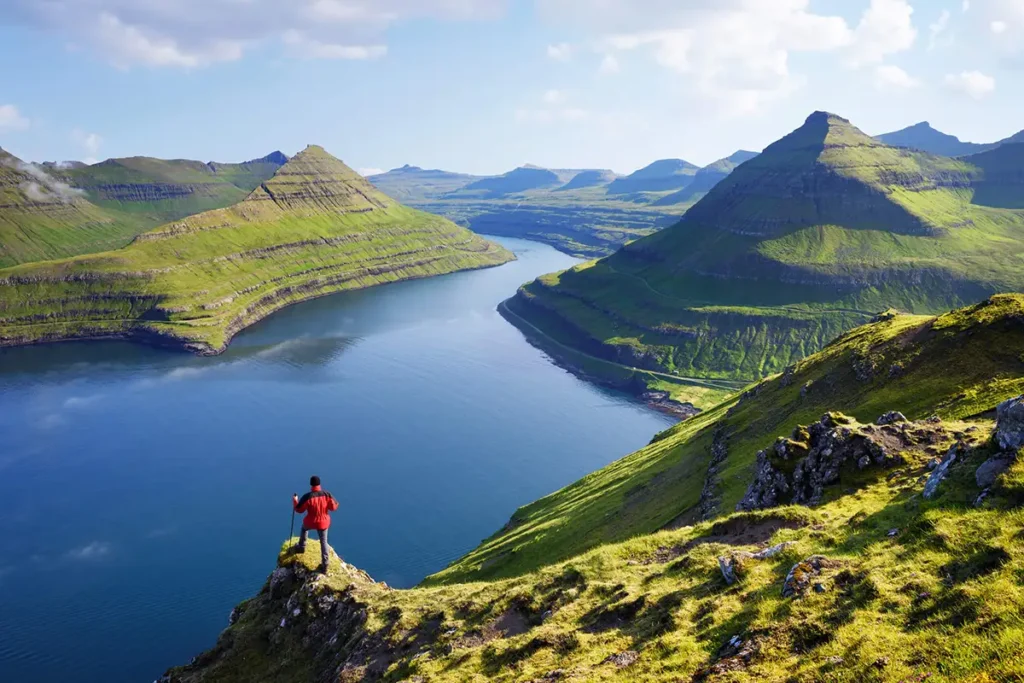 Experiencing-a-fjord-in-the-Faroe-Islands Hiker standing on a cliff overlooking a dramatic green fjord landscape in the Faroe Islands