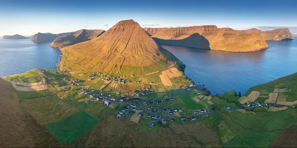 The-sun-sets-over-fjords-and-islands-in-the-Faroe-Islands Aerial view of a coastal village surrounded by steep green mountains and blue sea in the Faroe Islands