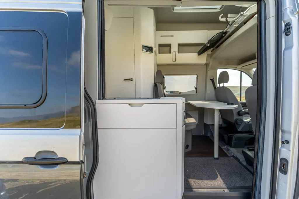 Campervan interior with seating area, table, and storage cabinets visible through side door