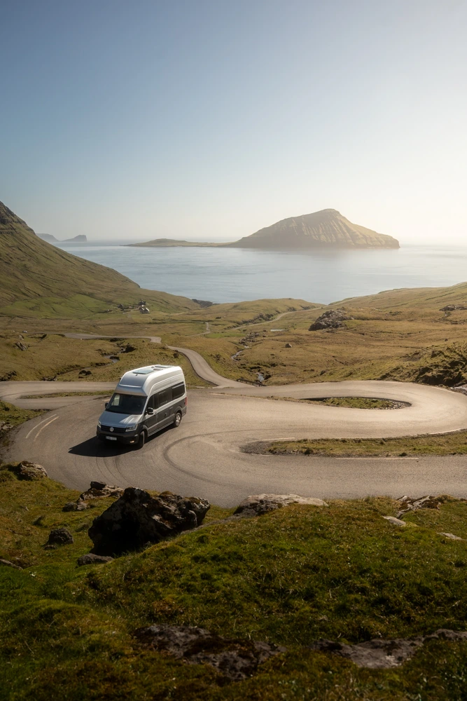 Campervan on a sunlit hairpin bend above Norðradalur, with Koltur across the sea.