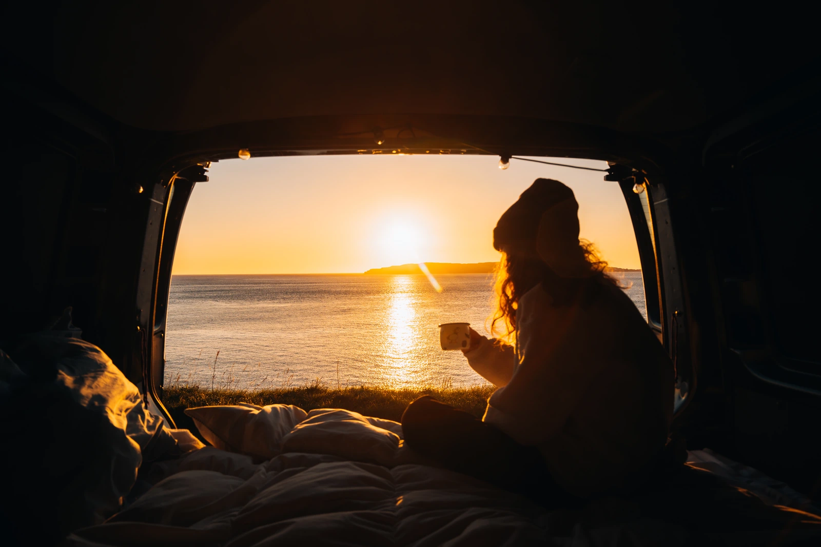 Traveller holding a mug inside a campervan while watching sunset over the sea.